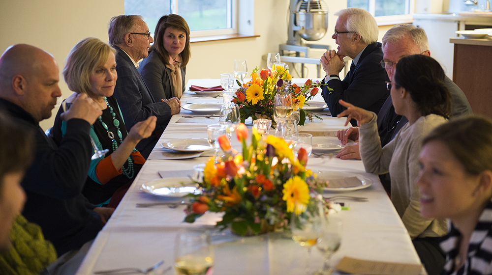 Multiple people gather at a table for a meal. The table is decorated with flowers and tableware.
